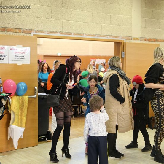 Eine Gruppe von Erwachsenen und Kindern versammelt sich in einem Saal. Einige Erwachsene tragen Kostüme. Eine Frau hält ein kleines Kind und ein Mädchen schaut zu ihnen. Bunte Luftballons sind an einer Wand befestigt. In der Ecke steht ein Mülleimer.