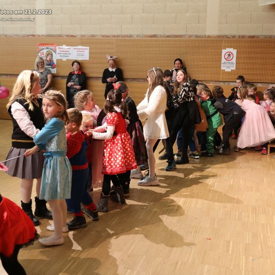 Eine Gruppe von Kindern in Kostümen, möglicherweise bei einer Schulveranstaltung, steht in einer Reihe in einem Raum. Eine Frau mit einem Stock steht vor ihnen. Einige Erwachsene stehen hinter ihnen und einige in der Nähe der Wand. Ein Schild befindet sich an der Wand. Das Datum 21.2.2023 steht oben links.