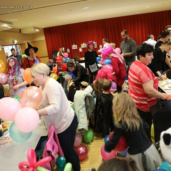 Eine Gruppe von Menschen feiert eine Karnevalsparty mit bunten Luftballons und Dekorationen. Einige tragen Kostüme, andere halten Luftballons. Der Hintergrund hat einen roten Vorhang.