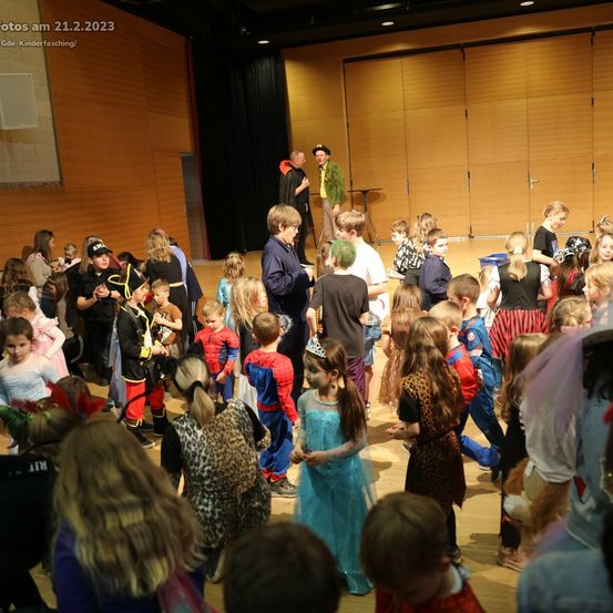 Eine Gruppe von Kindern in Kostümen auf einer Bühne, einige in Superhelden-Outfits, andere in Tierdrucken. Zwei Erwachsene in Kostümen sind auf der Bühne. Der Hintergrund besteht aus Holzwänden und einem schwarzen Vorhang.