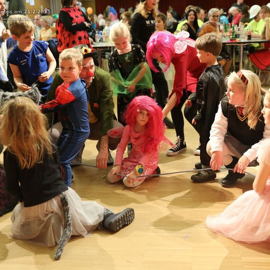 Kinder in Kostümen, darunter eine rosa Fee und ein Spiderman, spielen auf dem Boden, möglicherweise bei einer Halloween-Veranstaltung. Ein Clown mit roter Nase ist ebenfalls anwesend.