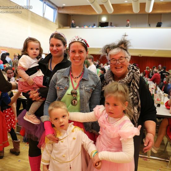 Eine Gruppe von Menschen in Kostümen posiert für ein Foto in einem Saal mit einem roten Vorhang im Hintergrund. Einige halten Kinder.