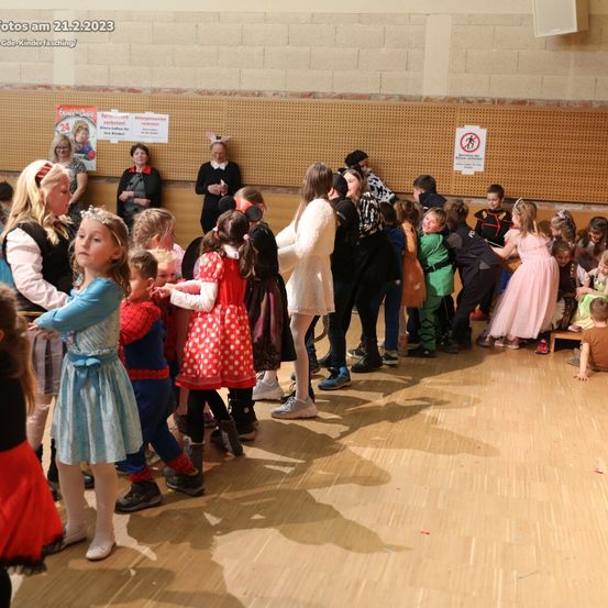 Kinder und Erwachsene versammeln sich am 21.2.2023 in einem Saal, einige in Kostümen. Einige Erwachsene stehen in der Nähe der Wand mit Schildern. Der Boden ist aus Holz.