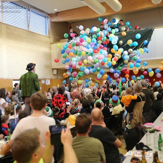 Eine Menschenmenge ist drinnen versammelt, einige in Kostümen, feiert. Ein Mann in Grün steht auf einem Balkon und lässt Ballons frei. Ein Kind in Orange und Schwarz hält eine Kamera. Tische sind mit Ballons und Getränken gedeckt.