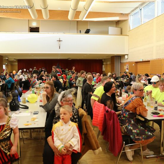 Eine große Gruppe von Menschen in verschiedenen Kostümen versammelt sich in einem Saal für einen festlichen Anlass, sitzt an Tischen und unterhält sich. Der Raum hat Holzböden, eine Galerie und einen roten Vorhang im Hintergrund.
