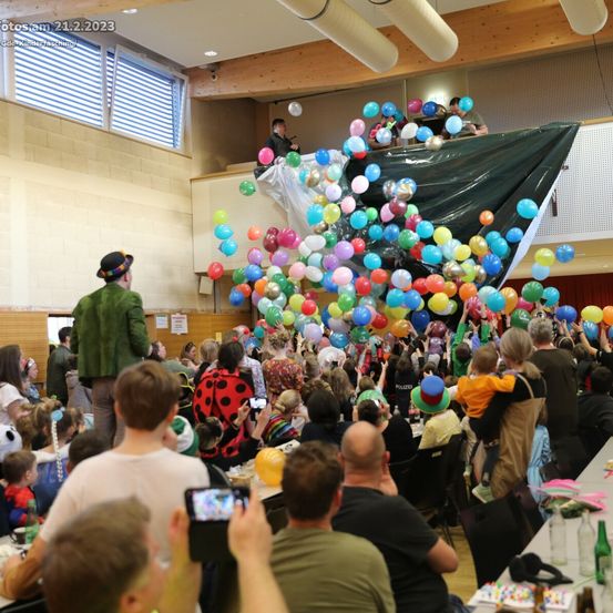 Eine große Menschenmenge versammelt sich in einem Innenbereich, möglicherweise für einen Karneval. Bunte Luftballons werden von oben freigelassen und schaffen eine festliche Atmosphäre. Einige Menschen tragen Kostüme und einige machen Fotos. Ein Mann in einem Hut steht in der Nähe der Ballons. Das Bild ist vom 21.2.2023.