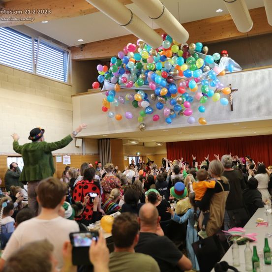 Eine festliche Zusammenkunft mit kostümierten Menschen, einem Mann in einem grünen Mantel und Hut, der die Menge anführt, schwebenden Ballons und einem Kreuz auf dem Balkon.