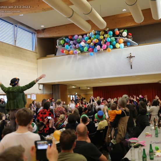 Eine große Menschenmenge ist in einem Raum versammelt, in dem Ballons von der Decke hängen. Einige Menschen heben ihre Hände, während andere Fotos machen. Der Raum hat eine Holzdecke und ein Kreuz an der Wand.