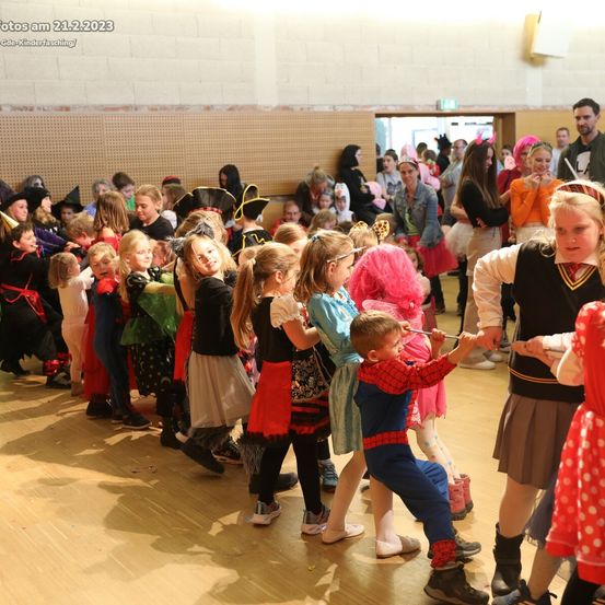 Eine Gruppe von Kindern in Kostümen bei einer Schulveranstaltung, die auf einem Holzboden tanzen und spielen. Ein junger Junge, verkleidet als Spiderman, interagiert mit einem Mädchen.