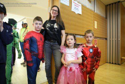 Eine Gruppe von Kindern in Superheldenkostümen posiert für ein Foto mit einer erwachsenen Frau in einem Flur mit Holzwänden und Schildern.