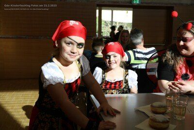 Drei junge Kinder in Piratenkostümen sitzen an einem Tisch mit Essen und Getränken. Dahinter sitzen eine Gruppe von Erwachsenen, von denen einer eine Marienkäfermaske trägt. Das Bild hat einen Wasserzeichen von Goede-Kinderfasching.