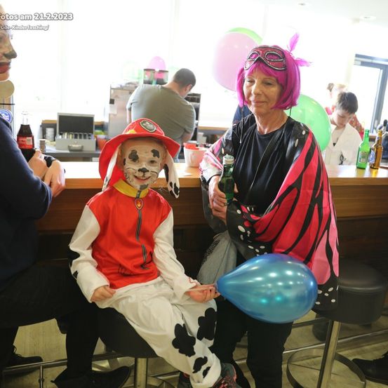 Ein junger Junge, verkleidet als Feuerwehrmann, steht neben einer Frau in einer Schmetterlingskostüm. Sie halten beide Luftballons. Im Hintergrund versammeln sich Leute um eine Theke.