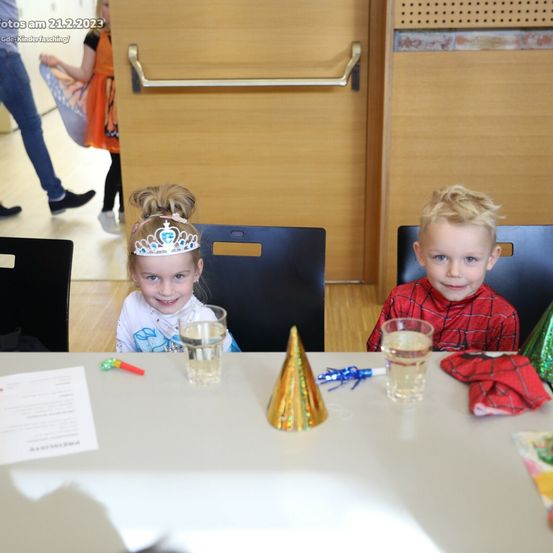 Zwei Kinder sitzen an einem Tisch mit Gläsern Wasser und Partyhüten. Das Mädchen trägt eine Krone und der Junge trägt ein rotes Spider-Man-Hemd. Hinter ihnen steht eine Tür.