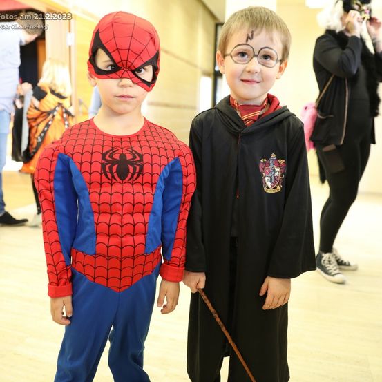 Zwei junge Jungen, einer als Spider-Man und der andere als Harry Potter verkleidet, stehen in einem Flur nebeneinander. Hinter ihnen sind andere Kinder in verschiedenen Kostümen zu sehen. Das Datumswasserzeichen ist 21.2.2023.