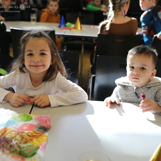 Ein Mädchen und ein Junge sitzen mit Luftballons und Bändern am Tisch, lächeln, in einem Raum mit anderen Kindern.