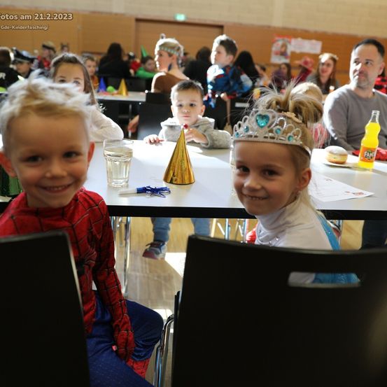 Eine Gruppe von Kindern und Erwachsenen sitzt in einem Saal an Tischen, vielleicht für eine Geburtstagsfeier. Ein Kind trägt ein Spiderman-Kostüm und ein anderes eine Krone. Auf dem Tisch stehen eine Fanta-Flasche und ein Donut.
