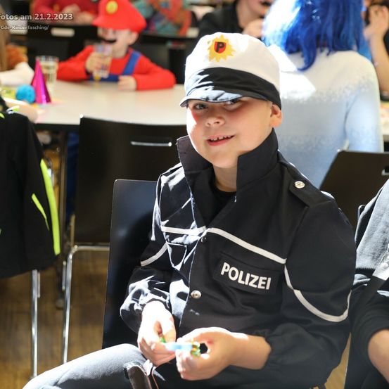 Ein Junge in einer Polizeiuniform sitzt auf einem Stuhl und lächelt. Hinter ihm sitzen eine Person mit blauen Haaren und ein Kind in Rot an einem Tisch. Das Bild wurde am 21. Februar 2023 aufgenommen.