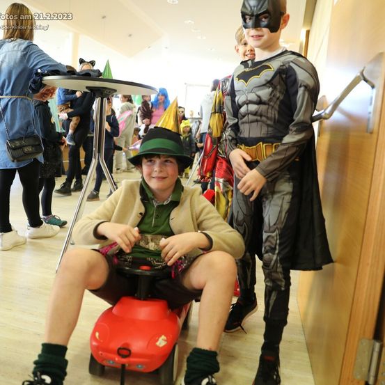 Zwei Jungen in Kostümen, einer als Batman und der andere in einem grünen Hut, posieren in einem Flur. Dahinter sitzt eine Person auf einem roten Spielzeugauto.