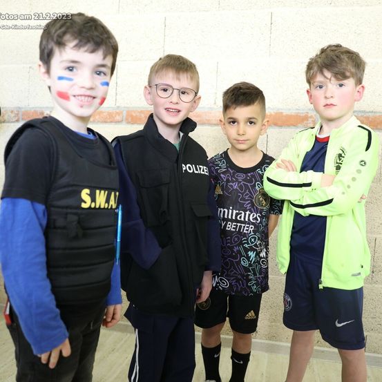 Vier Jungen stehen zusammen, zwei mit französischen Flaggen auf den Gesichtern. Einer trägt eine Polizeiweste, ein anderer ein schwarzes Shirt mit 'Emirates', einer eine blaue Jacke und ein anderer eine neonfarbene Jacke.