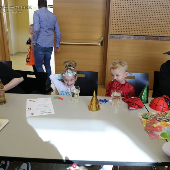 Zwei Kinder sitzen an einem Tisch in einem Partyraum mit Partyhüten, Getränken und Dekorationen. Ein Mann und ein Mädchen stehen im Hintergrund.