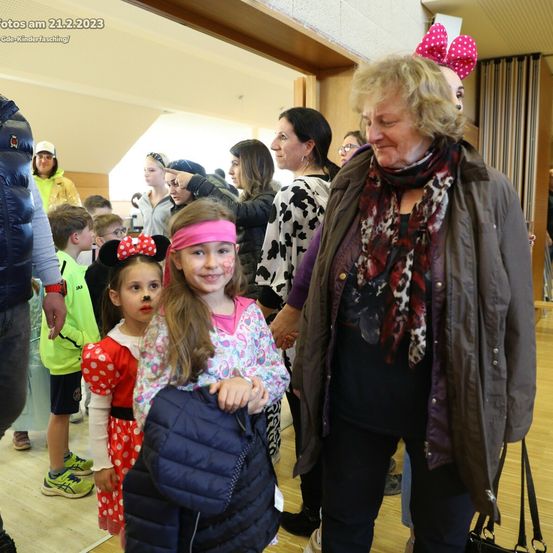 Eine Gruppe von Kindern in Kostümen, darunter Minnie Mouse, steht mit Erwachsenen in einem Raum. Das Datum auf dem Bild ist 21.2.2023.