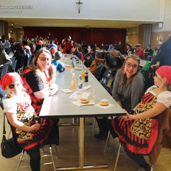 Eine Gruppe von Menschen in Kostümen an einem langen Tisch. Einige tragen Brillen und rote Kopftücher. Es gibt Teller, Gläser und Essen auf dem Tisch. Ein Kreuz ist an der Wand über dem Tisch angebracht.