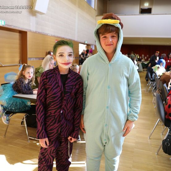 Zwei Kinder in Kostümen stehen in einem Saal zusammen. Ein Kind trägt ein Joker-Kostüm und das andere ein grünes Overall. Sie lächeln und posieren für ein Foto.