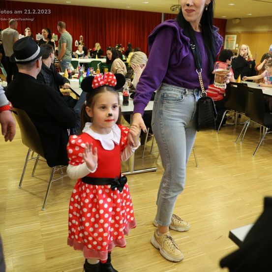 Ein kleines Mädchen, gekleidet wie Minnie Maus, wird von einer Frau in einem Raum mit Holzboden geführt. Andere Menschen sitzen an Tischen. Das Datum 21.2.2023 ist in der oberen linken Ecke sichtbar.