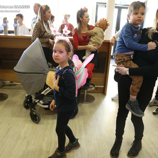 Eine Gruppe von Menschen, einschließlich Kindern und Erwachsenen, befindet sich in einem Raum mit einer hölzernen Theke. Ein junges Mädchen in einem Feen-Kostüm steht neben einem Kinderwagen. Eine Frau hält ein Baby an ihrem Arm.