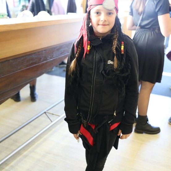 Ein junges Mädchen in einem Piratenhut und einer schwarzen Jacke mit rotem Saum steht drinnen. Hinter ihr ist ein verschwommener Blick auf eine Holztheke und eine Frau in einem schwarzen Kleid.