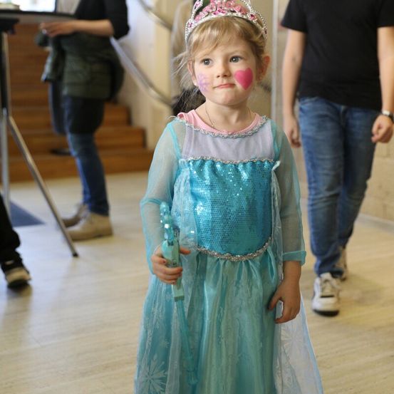 Ein kleines Mädchen, verkleidet als Elsa aus Frozen, hält einen Zauberstab. Sie hat Gesichtsbemalung und steht mit zwei Erwachsenen hinter sich auf einem Holzboden.