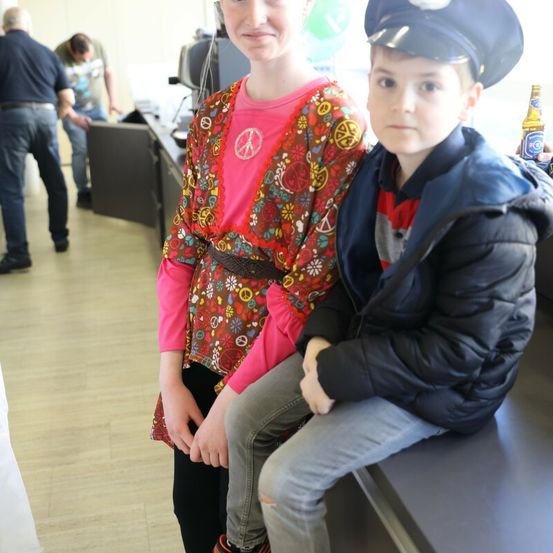 Zwei Kinder, ein Mädchen in einem rosa Kleid und ein Junge in einer Polizeiuniform, posieren für ein Foto in einer Innenumgebung. Hinter ihnen beugt sich ein Mann über einen Schreibtisch.