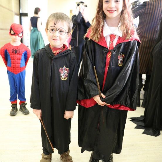 Zwei Kinder in Harry-Potter-Roben, eines mit einem Zauberstab, stehen neben einem Kind, das als Spiderman verkleidet ist. Dahinter stehen Menschen in verschiedenen Kostümen, wahrscheinlich bei einer thematischen Veranstaltung.
