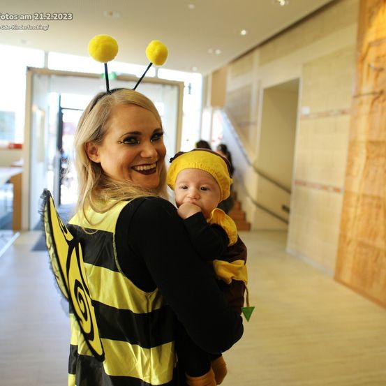 Eine Frau in Bienenkostüm hält ein Baby in Bienenkostüm und lächelt in einem Flur.