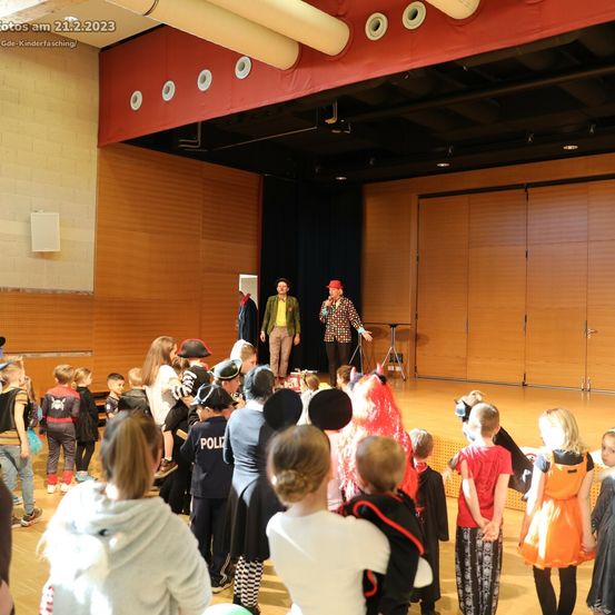 Eine Gruppe von Kindern in Kostümen beobachtet eine Aufführung auf der Bühne mit zwei Erwachsenen. Die Bühne hat Holzwände und einen roten Vorhang. Das Datum 21.2.2023 ist in der oberen linken Ecke sichtbar.