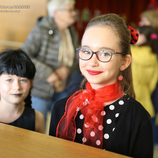 Ein Mädchen in einem Punktehemd und einem roten Schal sitzt an einem Tisch mit einem anderen Mädchen. Eine ältere Person mit Brille steht im Hintergrund. Das Bild ist vom 21. Februar 2023.
