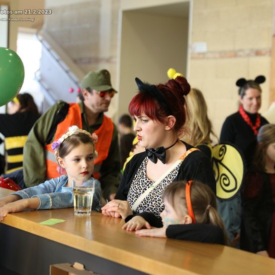 Eine Frau mit roten Haaren und einem Bienenkostüm sitzt mit zwei Kindern an einem Tisch. Die Kinder tragen Bienenkostüme. Ein Mann in Uniform steht hinter ihnen. Im Hintergrund befindet sich ein grüner Ballon.