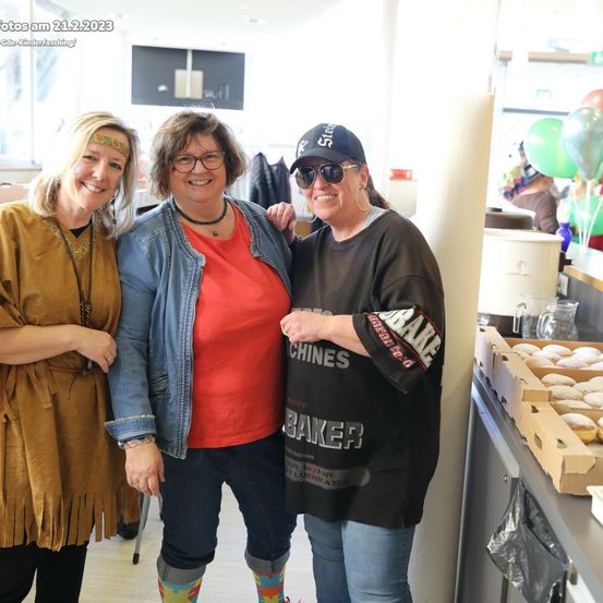 Drei Frauen in Freizeitkleidung stehen gemeinsam in einem Raum, lächeln und posieren für ein Foto. Eine hält einen Donut. Im Hintergrund sind bunte Luftballons und ein Tisch mit Gebäck zu sehen.