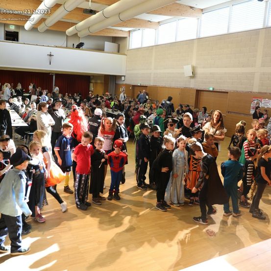 Eine große Menschenmenge von Kindern in Kostümen versammelt sich drinnen, wahrscheinlich für eine Halloween-Veranstaltung, mit Erwachsenen in der Nähe, einige tragen Hüte.