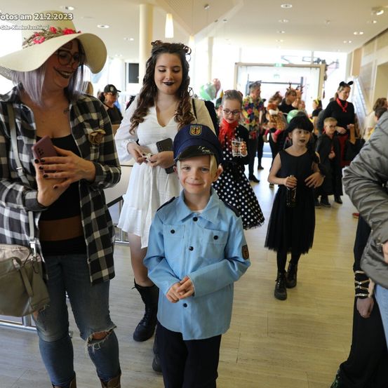 Mehrere Erwachsene und Kinder in Kostümen gehen in einem Flur. Ein junger Junge in einem blauen Uniform steht davor.