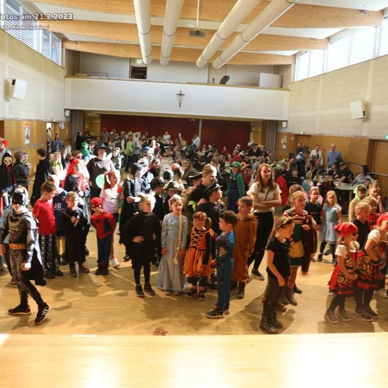 Eine große Gruppe von Kindern in verschiedenen Kostümen versammelt sich in einem Saal, einige stehen auf dem Holzboden. Der Saal hat Holzbalken und Glasfenster.