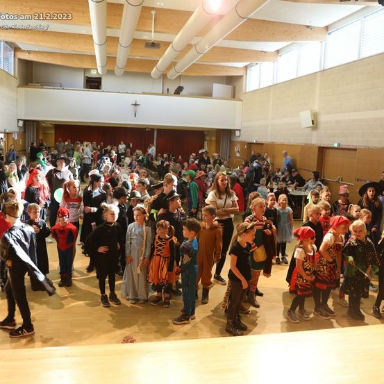Eine große Gruppe von Kindern in Kostümen ist in einer Halle versammelt, wahrscheinlich für eine Halloween-Veranstaltung. Der Saal hat Holzböden und eine hohe Decke mit Lichtern.