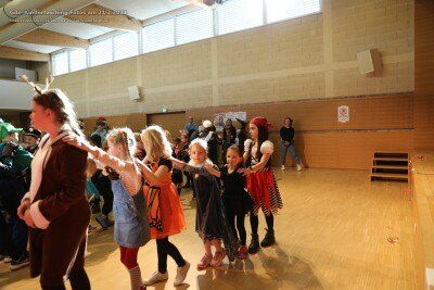 In einer Turnhalle tanzen eine Gruppe von Kindern in Kostümen, darunter ein Pirat, mit einem Erwachsenen. Die Turnhalle hat Holzböden und große Fenster.