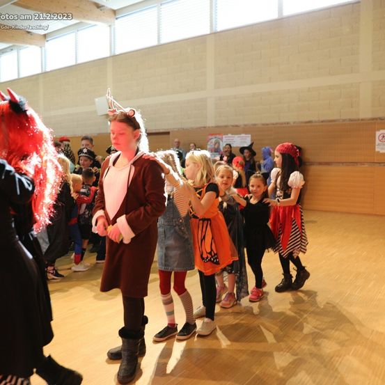 Eine Gruppe von Kindern in Kostümen, möglicherweise für ein Halloween-Event, steht in einem Raum. Sie tragen verschiedene Kostüme und einige von ihnen halten sich an den Händen. Der Raum hat einen Holzboden und weiße Wände.