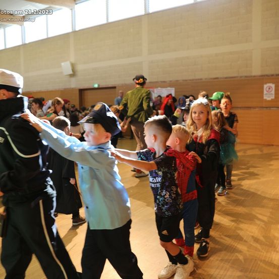 Drinnen tanzen Kinder in Kostümen, einige mit Erwachsenen. Ein Junge im Spiderman-Shirt hält ein Kind. Erwachsene in Uniformen stehen in der Nähe.