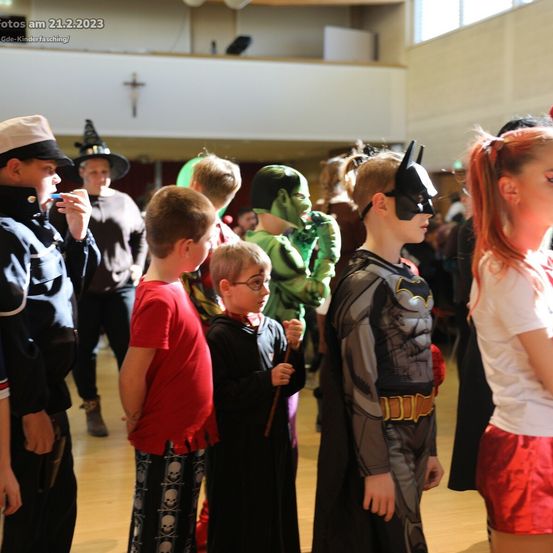 Eine Gruppe von Kindern in verschiedenen Kostümen steht in einem Saal. Ein Kind ist als Batman verkleidet, andere als Hexe, Hulk und Pirat. Sie scheinen an einer Kostümveranstaltung teilzunehmen.