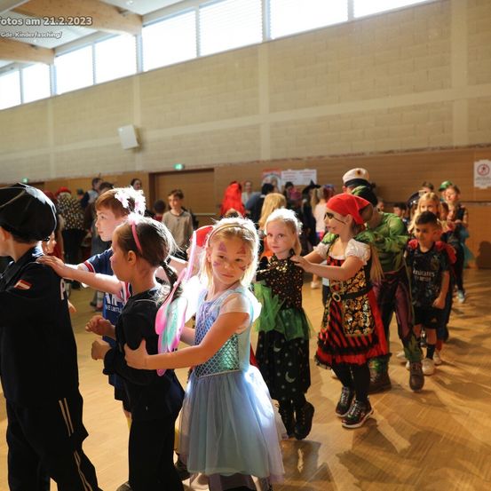 Gruppe von Kindern in Kostümen tanzt in einer Halle. Einige haben Gesichtsbemalung. Das Datum 21.2.2023 ist oben links sichtbar.