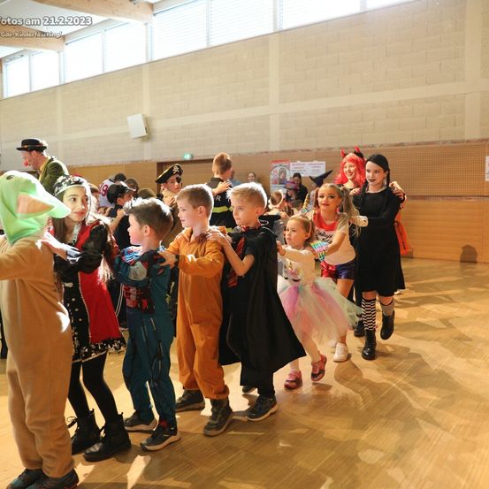 Eine Gruppe von Kindern in verschiedenen Kostümen versammelt sich in einem Raum. Sie stehen im Kreis und einige halten sich an den Händen. Sie tanzen wahrscheinlich. Der Raum hat einen Holzboden und eine beige Wand. In der oberen linken Ecke befindet sich ein Wasserzeichen.