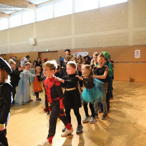 Eine Gruppe von Kindern in Kostümen tanzt am 21.2.2023 in einer Halle. Einige tragen Superheldenkostüme, andere haben Feenflügel.