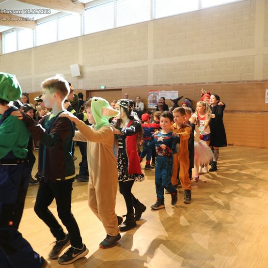 Eine Gruppe von Kindern in Kostümen tanzt in einer Halle mit Holzböden und Ziegelwänden. Im oberen linken Eck ist das Datum 21.2.2023 zu sehen.